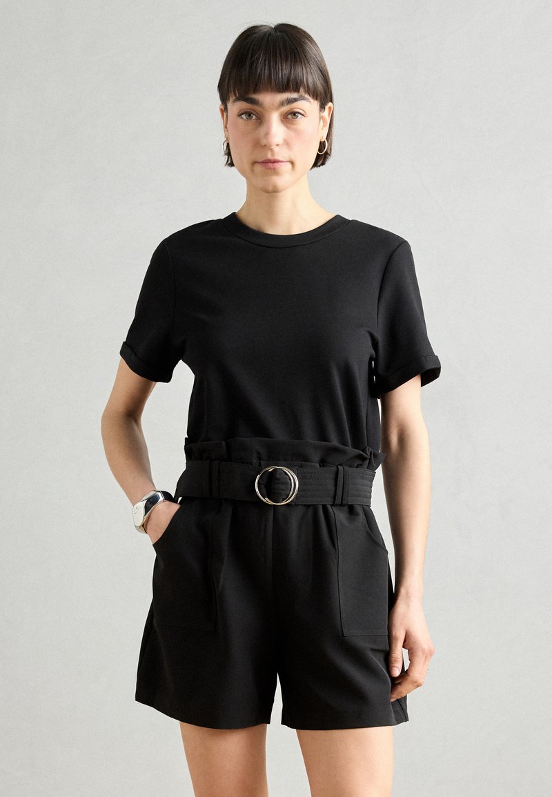 Haut noir à manches courtes associé à un short noir taille haute avec une ceinture en tissu à boucle circulaire et deux poches avant.