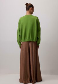 Zelen pulover iz pletenine z ohlapnim krojem in spuščenimi rameni, kombiniran z rjavimi hlačami širokih nogavic, ki poudarjajo gladke teksture.