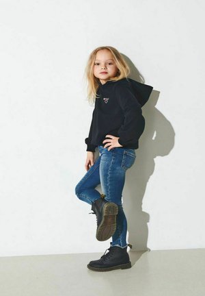 Sweat à capuche noir avec logo, jean slim bleu, et bottes noires à lacets avec semelles texturées. Debout devant un fond blanc.