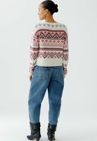 Gebreide trui in roomkleur met rode en zwarte geometrische patronen, gecombineerd met losse blauwe denim broeken en zwarte haklaarzen.