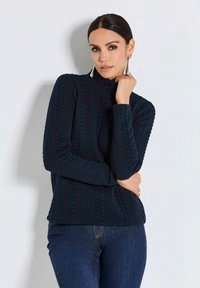 Dunkelblauer Strickpullover mit hohem Kragen, der ein strukturiertes Kabelmuster, lange Ärmel und einen lockeren Schnitt aufweist. Kombiniert mit Jeans.