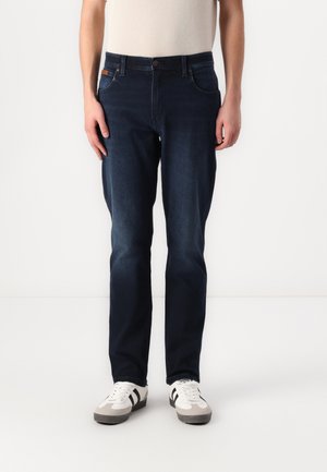Pantalones vaqueros de mezclilla azul oscuro con corte recto, diseño de cinco bolsillos, cierre de botón de metal y textura sutil. Combinados con zapatillas blancas y grises.