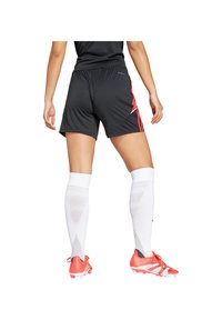 Persona in piedi di spalle, che indossa pantaloni corti neri con strisce rosse, calze bianche fino al ginocchio e scarpe da calcio arancioni brillanti.