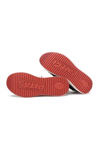 Black sneakers with red rubber soles, featuring deep grooves and the word "PITAS" embossed. Minimalist design with a low-top shape.