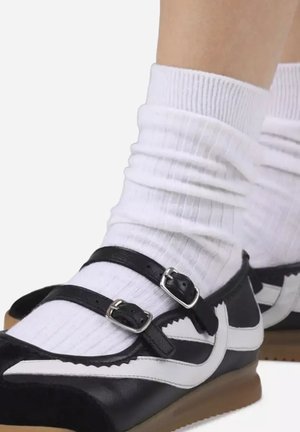 Pies con calcetines blancos acanalados y zapatos Mary Jane de cuero blanco y negro con doble hebilla sobre un fondo liso.