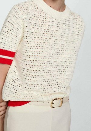 Personne portant un pull court en crochet blanc avec des rayures rouges sur les manches, un pantalon beige et une ceinture beige avec une boucle dorée.