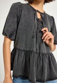 Kvinde justerer slips på sort, falmet denimbluse med puffærmer og samlede kanter, parret med blå jeans.