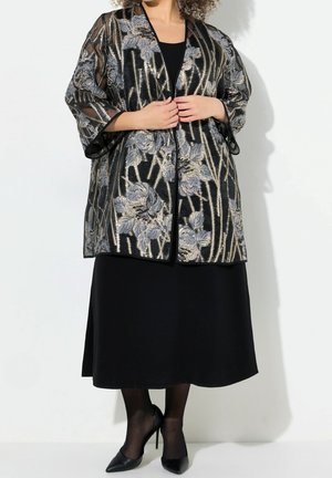 Femme portant une robe noire avec une veste noire transparente à motif floral métallique et des talons noirs.