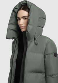 Giubbino imbottito verde oliva con colletto alto, caratterizzato da ampie sezioni trapuntate, una zip frontale e un logo circolare sulla manica.