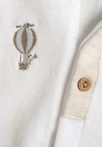 Chemise en tissu polaire blanc avec un design de montgolfière brodé beige. Comprend un bouton beige avec une surface texturée.