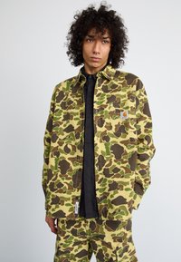 Camo-print overshirt in olive green, brown, and yellow hues; button-down design with a front pocket featuring a small logo patch.
