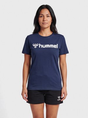 Vrouw met lang donker haar, die een donkerblauw hummel T-shirt en zwarte hummel shorts draagt, staand voor een effen lichte achtergrond.