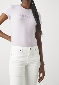 Světle fialové přiléhavé tričko s logem "ARMANI EXCHANGE" v tmavě fialové barvě, v kombinaci s bílými přiléhavými džíny. Hladká bavlněná látka, krátké rukávy.