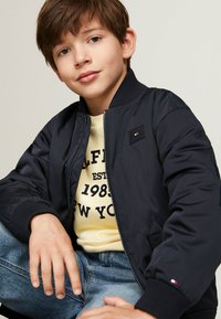 Giacca bomber navy con colletto e polsini a costine, caratterizzata da un piccolo logo. Indossata sopra una t-shirt grafica gialla e jeans azzurri chiari.
