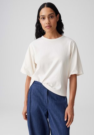 Donna con capelli lunghi e scuri, indossa una maglietta bianca a maniche corte e pantaloni blu a vita alta, in piedi davanti a uno sfondo semplice.