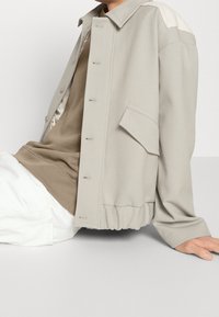 Chaqueta beige claro de corte relajado, con cierre de botones en el frente, bolsillo lateral y detalles texturizados en los hombros. Llevada sobre una blusa marrón.