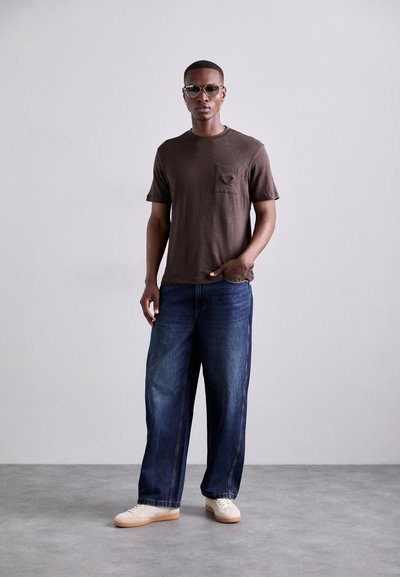 Brun kortärmad t-shirt med ficka, kombinerad med vida bl�å jeans och vita sneakers, modell som står mot en ljusgrå bakgrund.