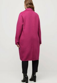 Lungo cappotto rosa con colletto, texture compatta e spacco posteriore singolo, abbinato a pantaloni neri aderenti e stivaletti lucidi alla caviglia.