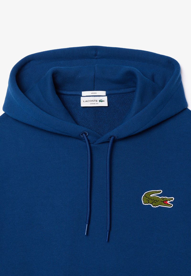 Sudadera con capucha azul hecha de algodón, con un corte holgado, cordones y un pequeño logo de cocodrilo verde en el pecho.