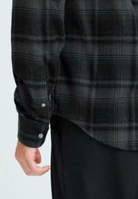 Black and gray plaid flannel shirt with buttoned cuffs, relaxed fit, rounded hem, and textured fabric, partially visible sleeve and hand.