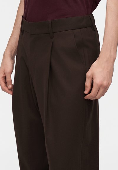 Homme portant un pantalon marron foncé sur mesure et une chemise bordeaux foncé, les mains détendues sur les côtés.
