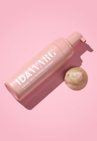 Bouteille rose de mousse autobronzante au cacao IDAWARG, avec un embout pompe. Produit mousseux brun clair à côté.