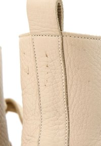 Gros plan sur la tige d'une botte en cuir beige avec des coutures visibles et de petites taches foncées sur la surface texturée.