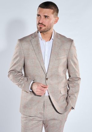 Mann im beigen karierten Anzug und weißem Hemd steht mit einer Hand in der Tasche und der anderen, die einen Jackenknopf richtet, seitlich schauend.