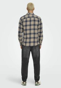 Karierte Langarmbluse in Blau, Braun und Creme. Aus einem Stoff mit weicher Textur gefertigt. Kombiniert mit dunklen Jeans und Sneakers.