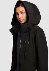 Black insulated jacket with a high collar and large hood; features zippers and a logo patch on the sleeve for added detail.