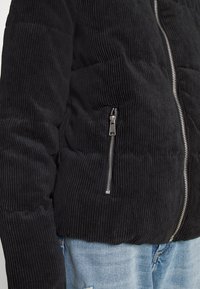 Svart kordflosjacka med vertikala ribbor, utrustad med en framzipper och sidofickor. Tyget har en strukturerad yta och en figurnära passform.