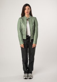 Giacca in pelle verde chiaro con colletto rialzato, tasche con zip e finitura testurizzata, abbinata a jeans scuri e sneaker.