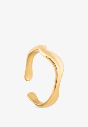 Frau mit nassen, dunklen Haaren berührt mit schlanken Fingern, die einen glatten, goldenen, gebogenen Ring am Mittelfinger tragen, ihre Lippen vor einem einfarbigen Hintergrund.