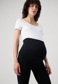 Leggings de maternité noirs avec une texture lisse et extensible, dotés d'une taille haute pour un meilleur maintien. Le haut est une chemise ajustée à manches courtes de couleur blanche avec un décolleté en scoop.
