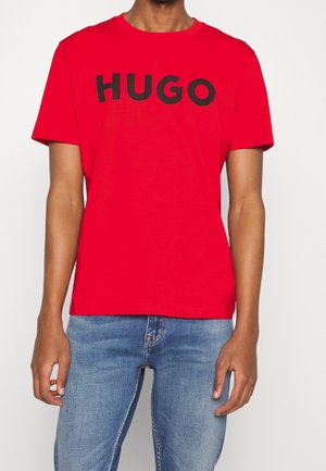 Man die een rood T-shirt met korte mouwen draagt met zwarte tekst "HUGO" en een blauwe spijkerbroek, staand tegen een effen witte achtergrond.