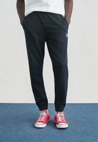 Μαύρο παντελόνι φόρμας με ελαστική μέση και πλαϊνές τσέπες, σε συνδυασμό με κόκκινα πάνινα παπούτσια με λευκές λαστιχένιες σόλες και κορδόνια.