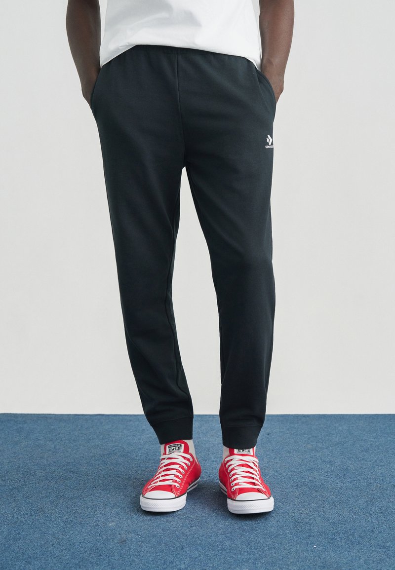 Μαύρο παντελόνι φόρμας με ελαστική μέση και πλαϊνές τσέπες, σε συνδυασμό με κόκκινα πάνινα παπούτσια με λευκές λαστιχένιες σόλες και κορδόνια.