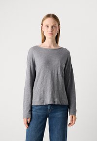 En ung kvinne med rett blondt hår har på seg en løs grå langermet skjorte og blå jeans, stående mot en enkel hvit bakgrunn.