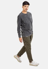 Man walking, wearing a faded black long-sleeve shirt, olive cargo pants, and white sneakers against a plain white background.