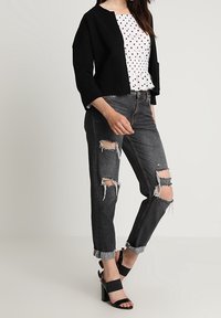 Veste noire cropped, blouse blanche à pois, jeans noirs déchirés avec ourlet effiloché et sandales à talons noires, affichant un style décontracté moderne.