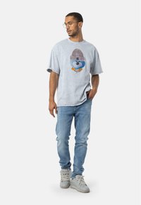 T-shirt oversize di colore azzurro con grafiche di un personaggio dei cartoni animati che indossa un cappello di maglia, abbinato a jeans azzurri strappati e sneakers grigie.