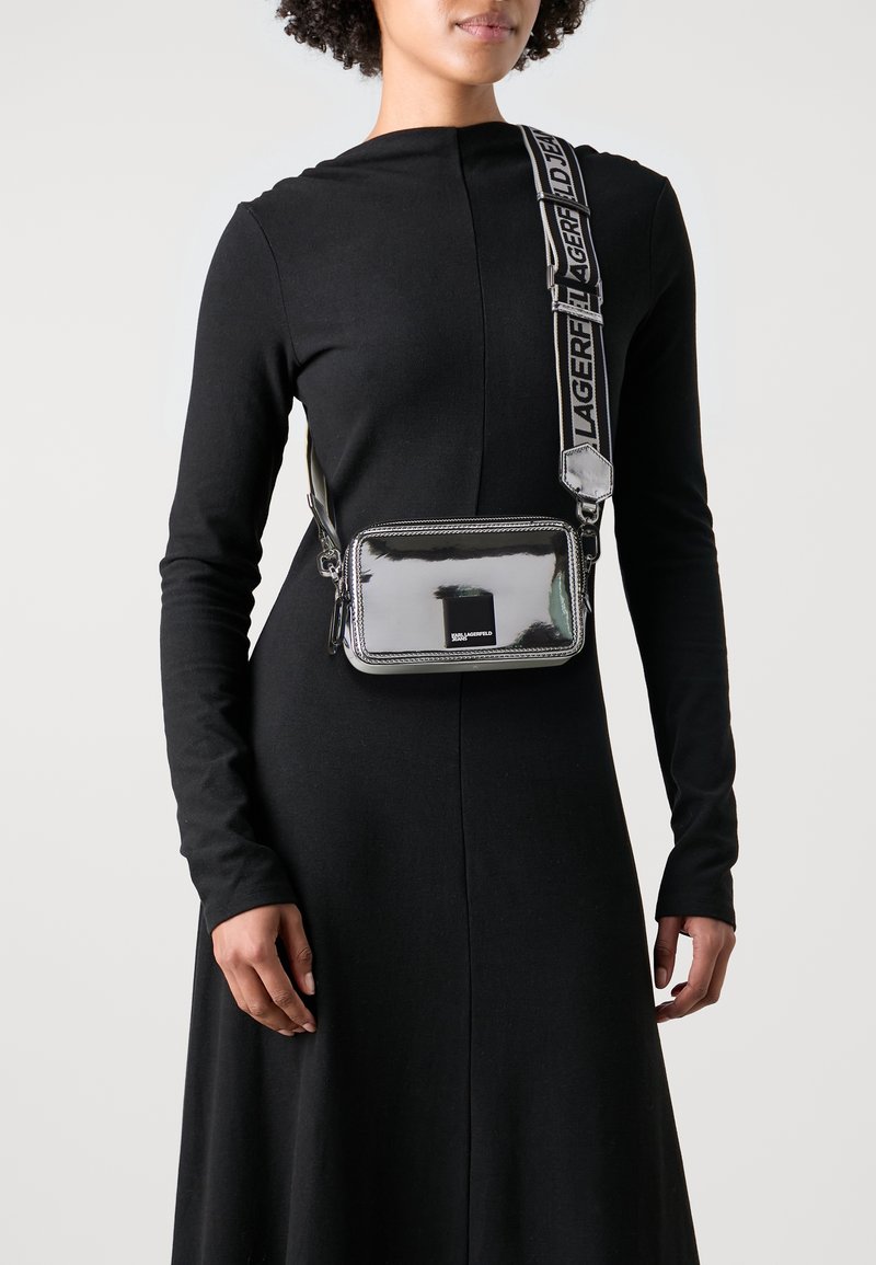 Borsa a tracolla argentata con finitura lucida, forma rettangolare, logo nero e tracolla con il marchio. Superficie liscia e dettaglio con zip.