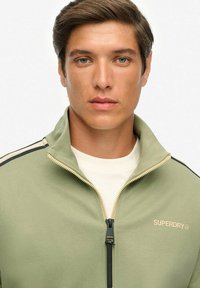 Chaqueta verde claro con cremallera y rayas negras, hecha de un material suave, con un cuello alto y la marca "SUPERDRY" en beige en el pecho.