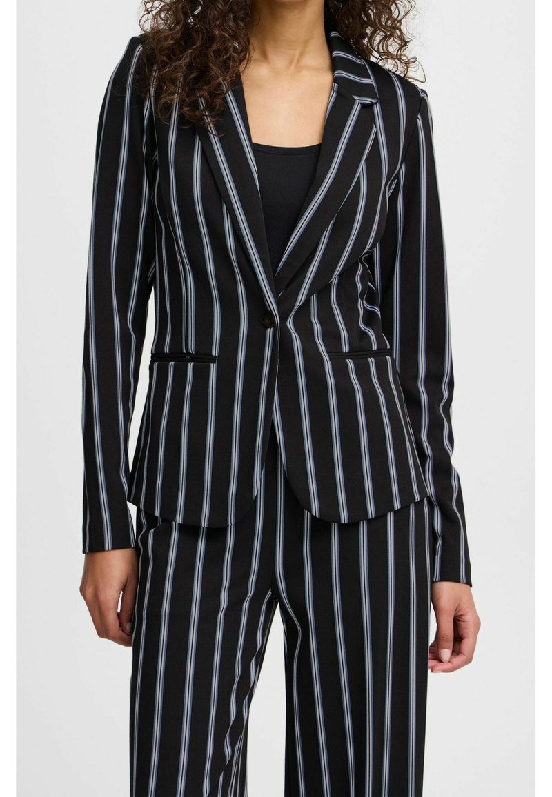 Femme portant un blazer noir et un pantalon à fines rayures verticales blanches, associé à un haut noir uni en dessous.