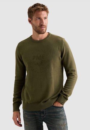 Man with short hair and beard wearing an olive green sweater with embossed text and wings, and blue distressed jeans, looking to the side.