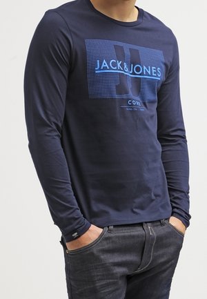 Man draagt een marineblauw Jack & Jones T-shirt met lange mouwen en heeft zijn handen in de zakken van een donkere spijkerbroek tegen een effen achtergrond.