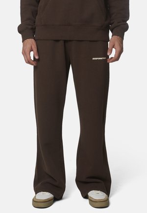 Persoon draagt donkerbruine wijdvallende sweatpants met witte tekst op het dijbeen en witte sneakers met beige zolen.