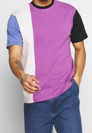Homme portant un t-shirt à manches courtes color block avec des sections violettes, noires, bleues et blanches, associé à un jean bleu et un bracelet argenté.
