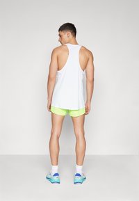 Witte mouwloze sporttanktop met een lage rug. Gecombineerd met felneon groene shorts en sneakers met blauwe accenten.