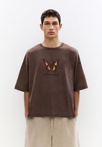 T-shirt oversize marrone con grafica di una farfalla in arancione e nero, con scollatura rotonda e dettagli delle cuciture in vista.